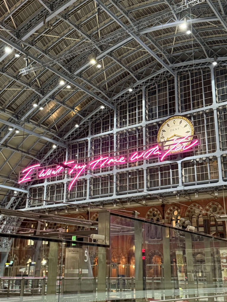 The vast steel-framed ceiling of St Pancras station in London, featuring a large clock and a prominent pink neon sign that reads,