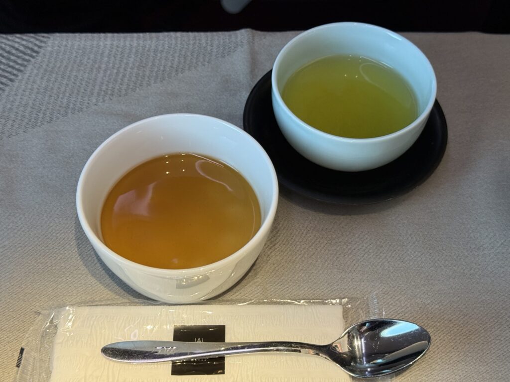 テーブルの上に置かれた、白い器に入った琥珀色のデザートと緑茶。手前にはJALのロゴが入ったナプキンとスプーンが添えられている。