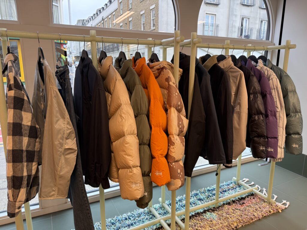 Inside a clothing store. Various types of outerwear, such as down jackets and plaid shirts, are hung in a row on a uniquely designed light green rack.
