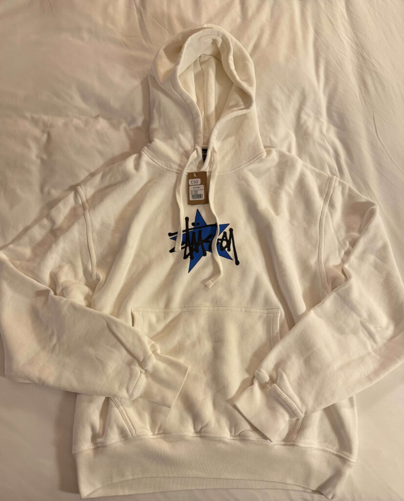 A white Stüssy hoodie with a blue star and black graffiti logo on the chest, laid out flat on a bed.