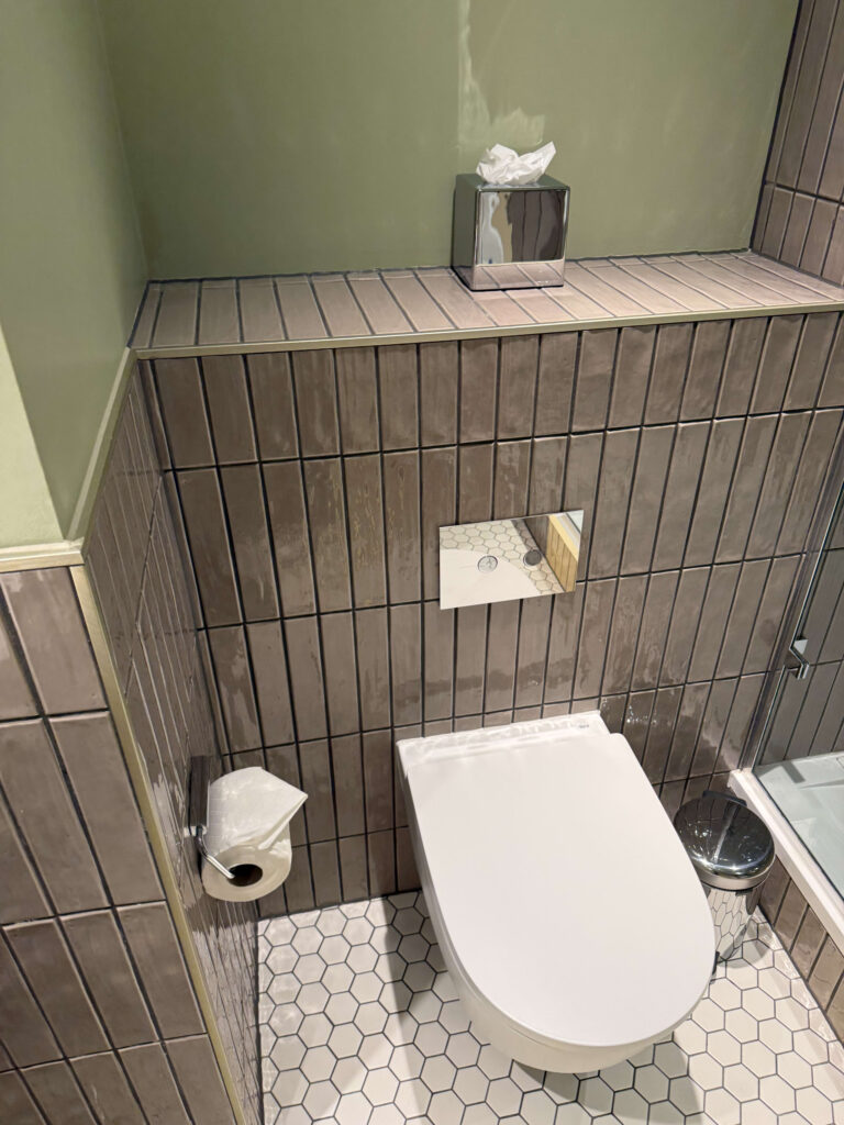 A toilet in a bathroom with olive green walls, brown vertical tiles, and a white hexagonal tile floor.