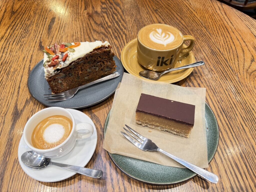 A slice of carrot cake, a caramel slice, and two cups of coffee, including a latte with art, are arranged on a wooden cafe table.