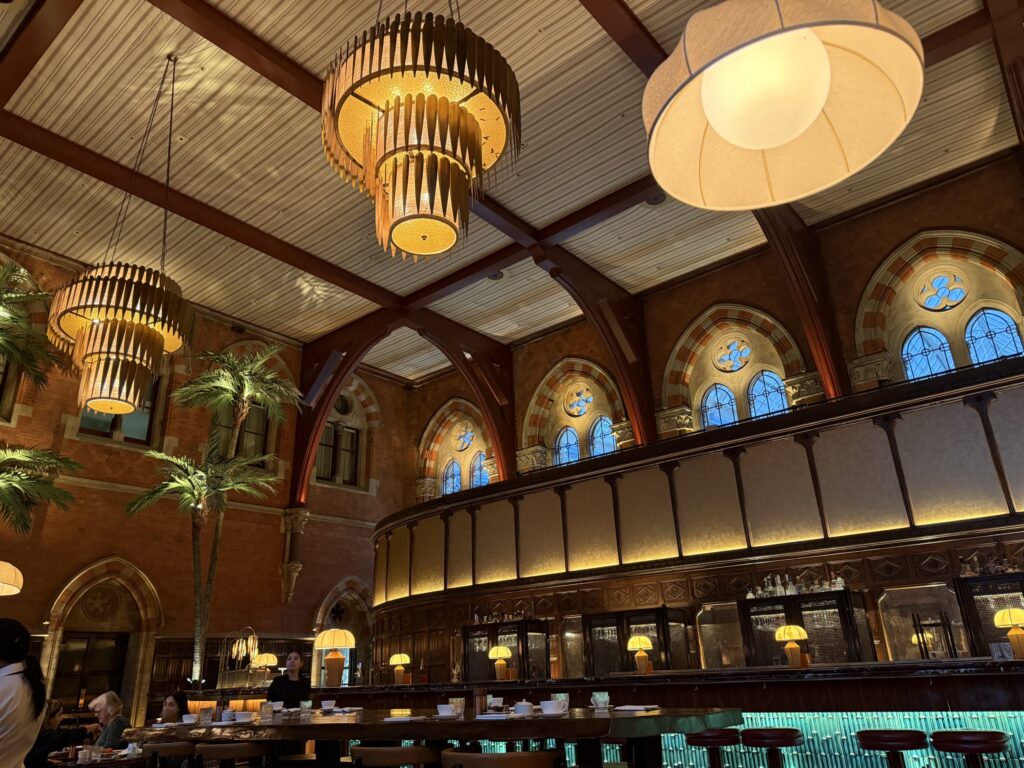 The interior of a grand restaurant with high ceilings, Gothic arched windows, and large modern chandeliers hanging over a long central bar counter.