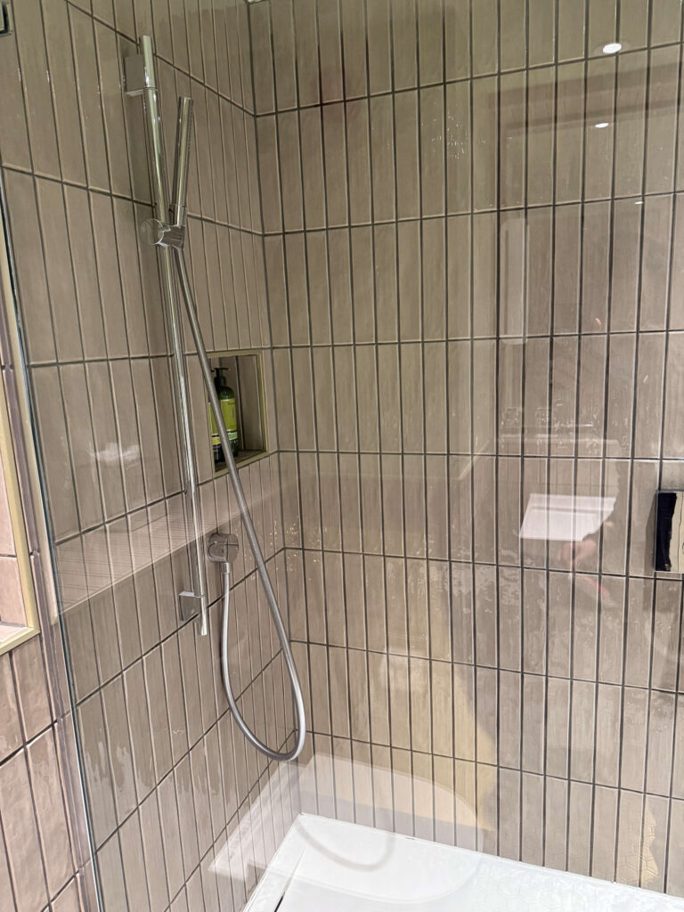 縦長のグレージュタイルが貼られたモダンなシャワールーム。クローム製のハンドシャワーと、壁のニッチに置かれたロクシタンのアメニティが見える。