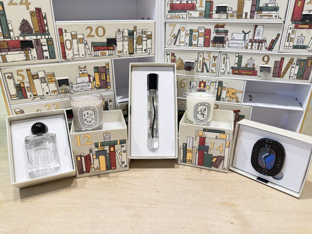 木製のテーブルの上に置かれたディプティックのアドベントカレンダー。11日から15日までの引き出しが開けられ、中から出てきたミニキャンドル2つ、香水2種類、ホリデーオーナメントが手前に並べられている。