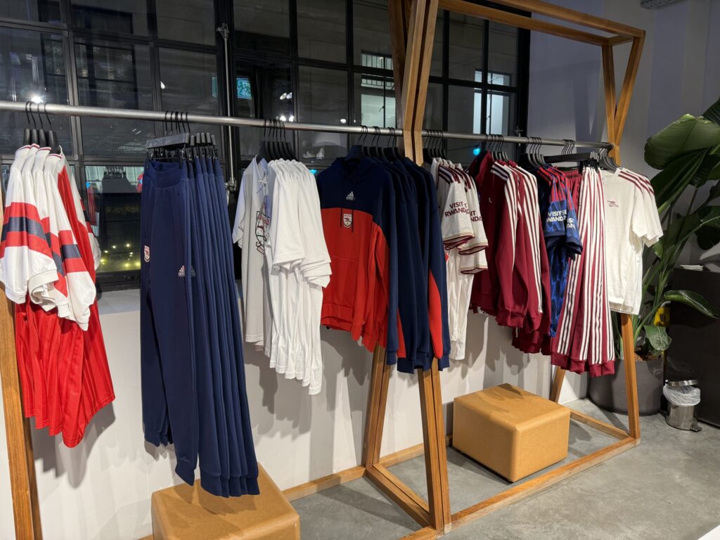 A wooden clothing rack inside a store, displaying a collection of Arsenal football club apparel, including jerseys and T-shirts.