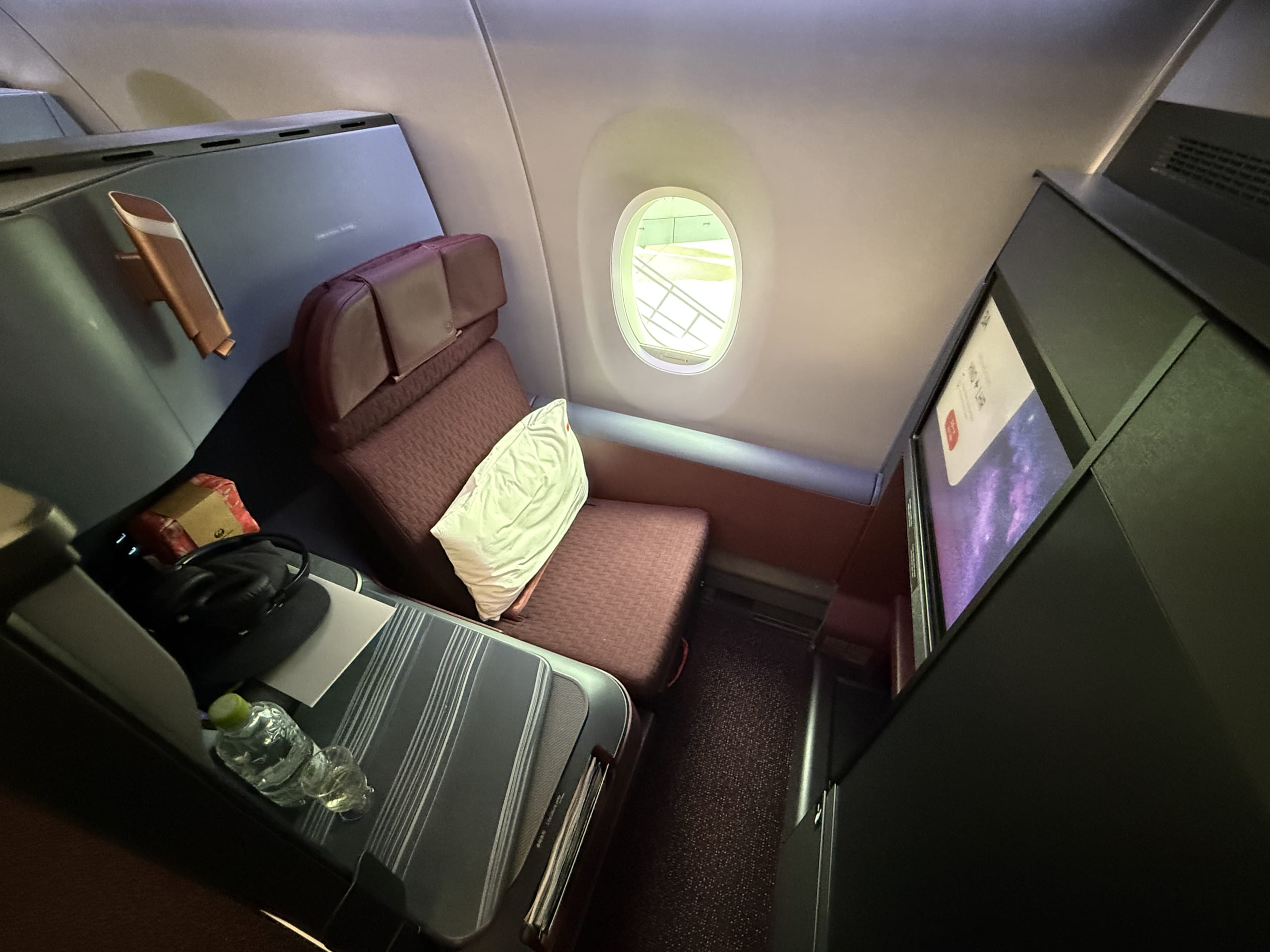 A private, suite-style business class seat on a JAL Airbus A350-1000. A wine-red seat with a white pillow is visible, along with a window, side table, and a large monitor.