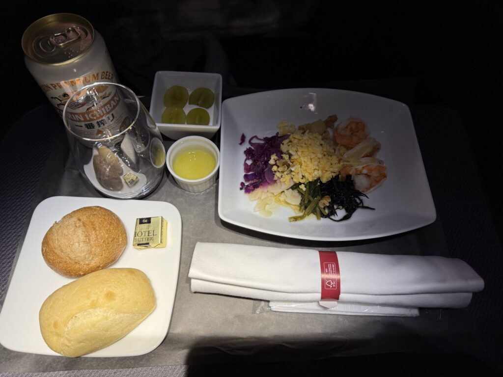 飛行機の座席テーブルに置かれた「フミコの洋食」の機内食。カラフルなサラダ、パン、ブドウ、そしてキリン一番搾りの缶ビールが並んでいる。