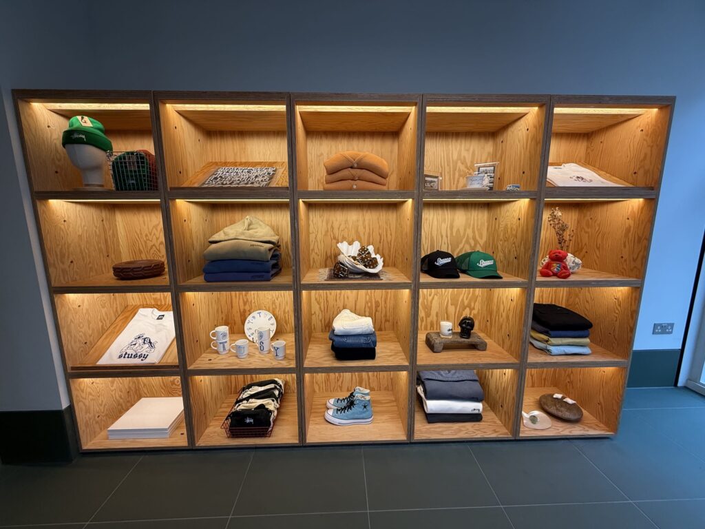 A wooden display shelf with multiple lit-up cubbies, showcasing various Stüssy brand merchandise including apparel like T-shirts and hoodies, hats, Converse sneakers, and other accessories.