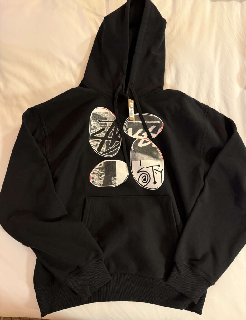 A black Stüssy x David Carson collaboration hoodie spread out on a white sheet, featuring an abstract collage graphic on the chest.