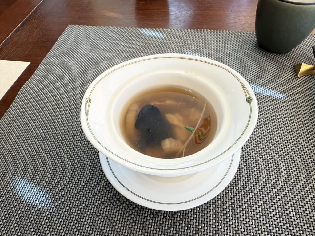 An amber-colored steamed soup with mountain delicacies and buckwheat groats served in an elegant white bowl. It is placed on a grey placemat on a wooden table.