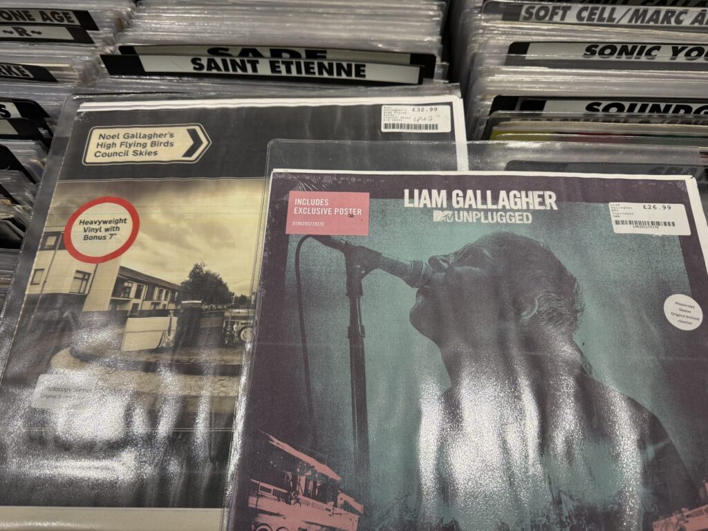 Vinyl records of Noel Gallagher's album 'Council Skies' and Liam Gallagher's album 'MTV Unplugged' side by side in a record store rack.