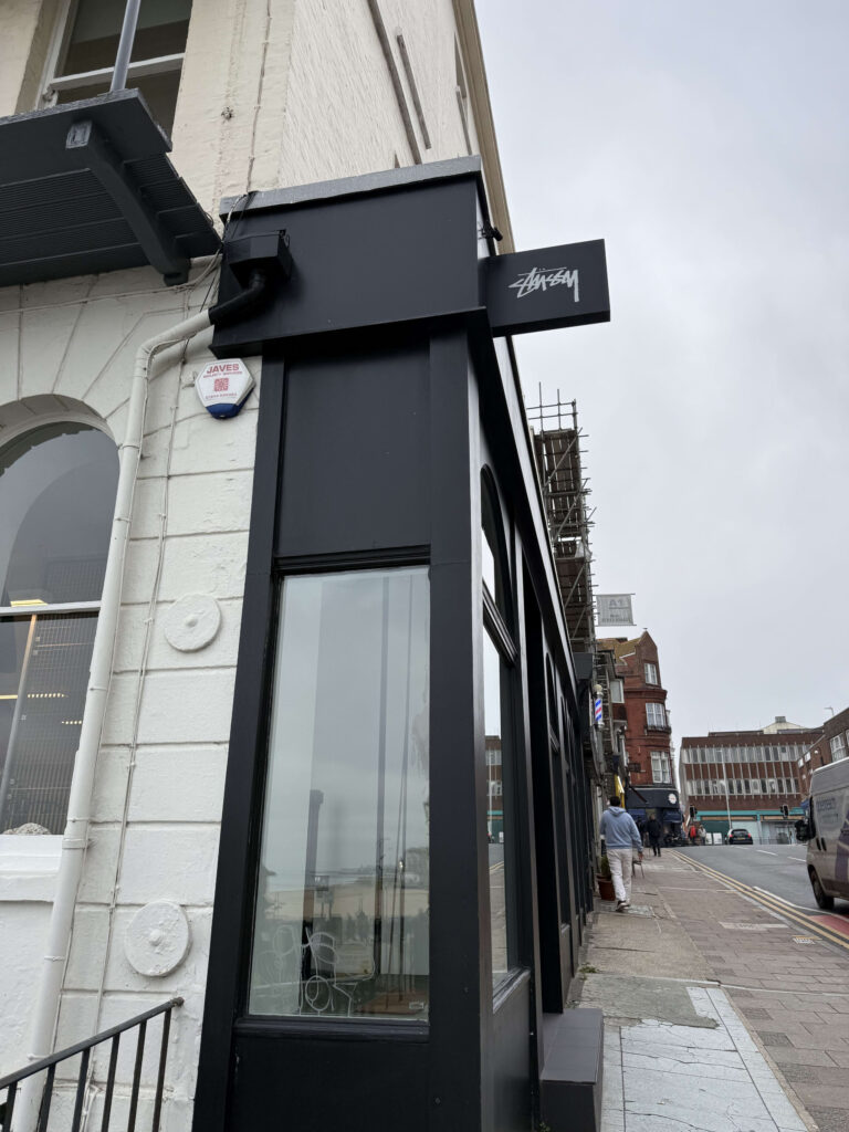 イギリスのマーゲートにあるStussyの店舗。白い壁の建物の角に、黒いファサードと白いロゴが入った看板が突き出している。