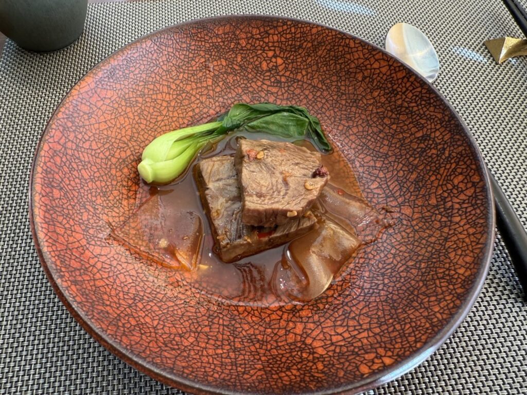 Braised beef cheek in a spicy Sichuan-style sauce, garnished with bok choy and served in an orange bowl with a crackle glaze pattern.
