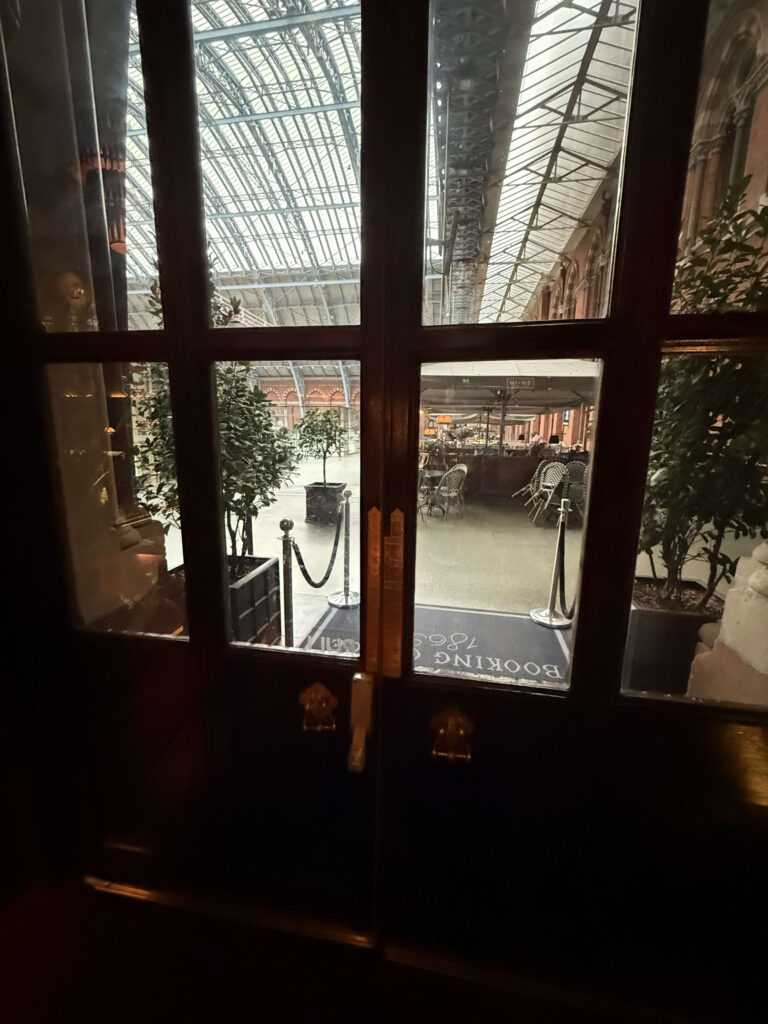 View through the glass panes of a dark wooden door looking out onto the bright concourse of St. Pancras International station with its high, arched glass roof.