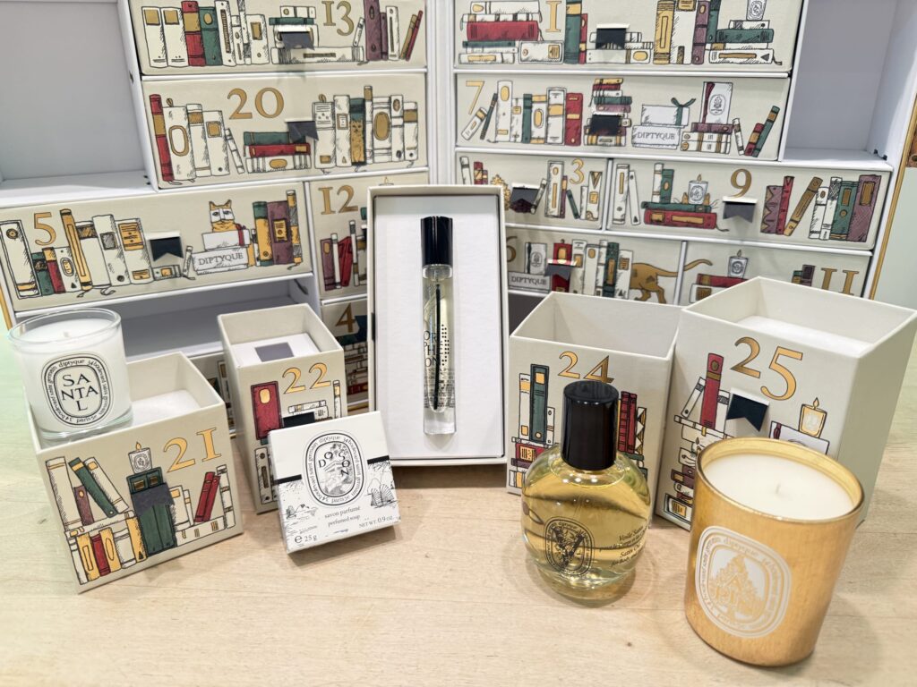 Diptyque advent calendar with open boxes for days 21 through 25, displaying various products like candles, perfume, soap, and body oil on a wooden table.