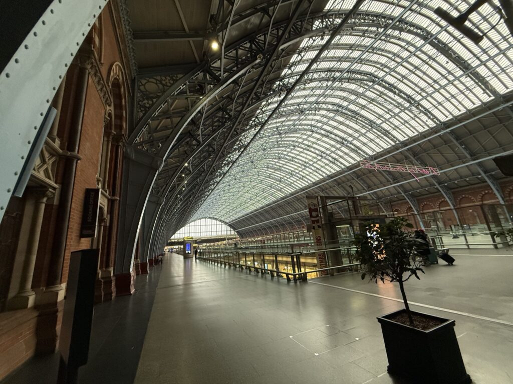 The interior of St Pancras International station in London. A vast, arched roof made of steel and glass covers the spacious platforms below. On the left, a red brick wall features a sign that reads 'BOOKING OFFICE.'