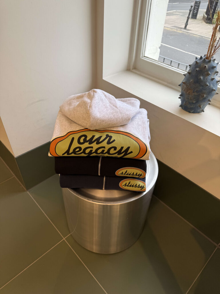 A stack of three Our Legacy and Stüssy collaboration hoodies on a cylindrical metallic stool. The top one is gray, with two navy hoodies underneath.