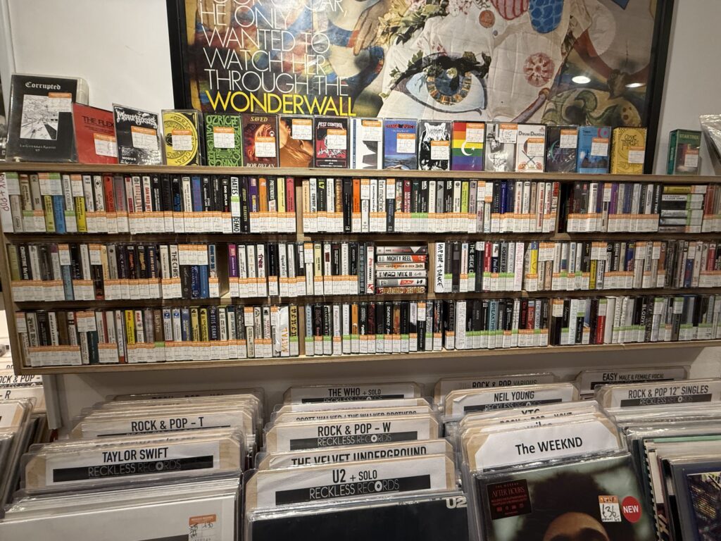 Shelves packed with cassette tapes in a record store. Below are bins of vinyl records from artists like Taylor Swift and U2.