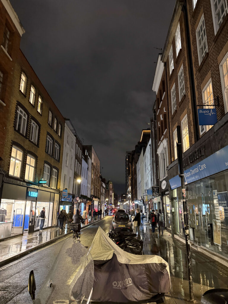 A nighttime view of Berwick Street in London. The pavement is wet, reflecting lights from the illuminated shops and windows of the brick buildings lining the street as people walk by.