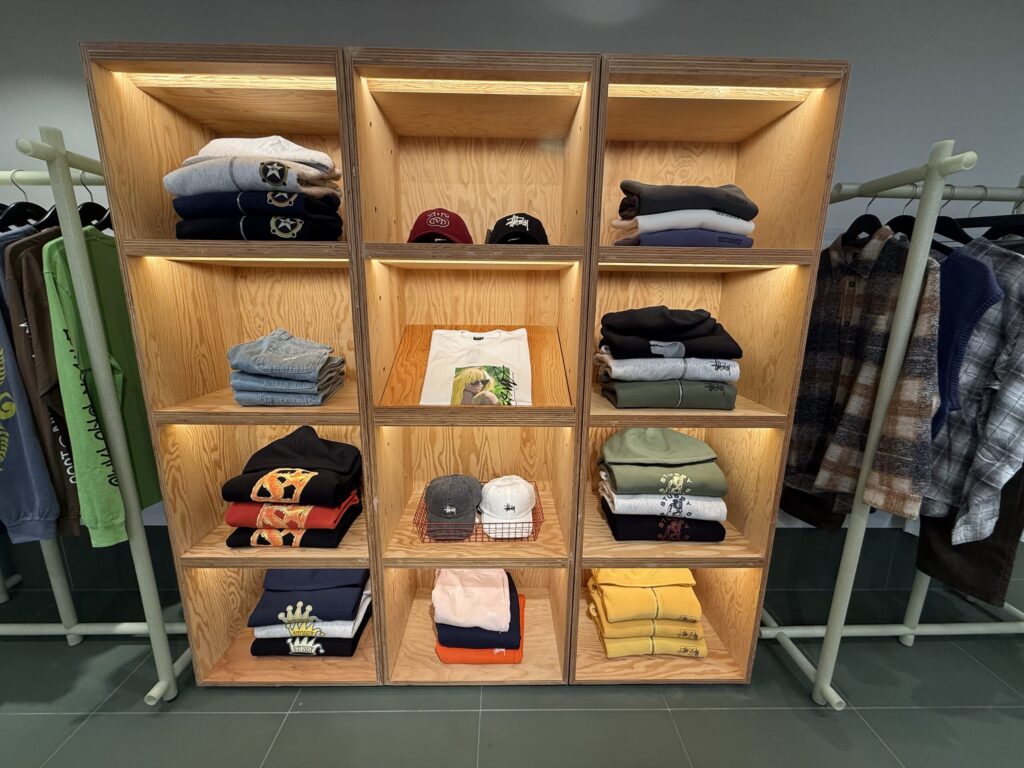 Inside a clothing store. Neatly folded sweatshirts, T-shirts, and caps are displayed on a wooden shelving unit illuminated by warm lighting. Clothing racks with hanging garments are visible on both sides.