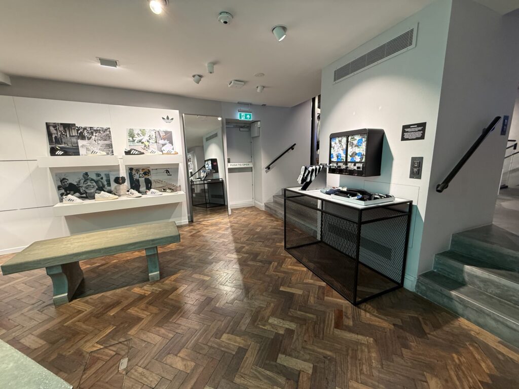 アディダスストアの店内。ヘリンボーン柄のフローリングの空間に、スニーカーが飾られた白い棚とベンチ、ステッカーの自動販売機が置かれている。