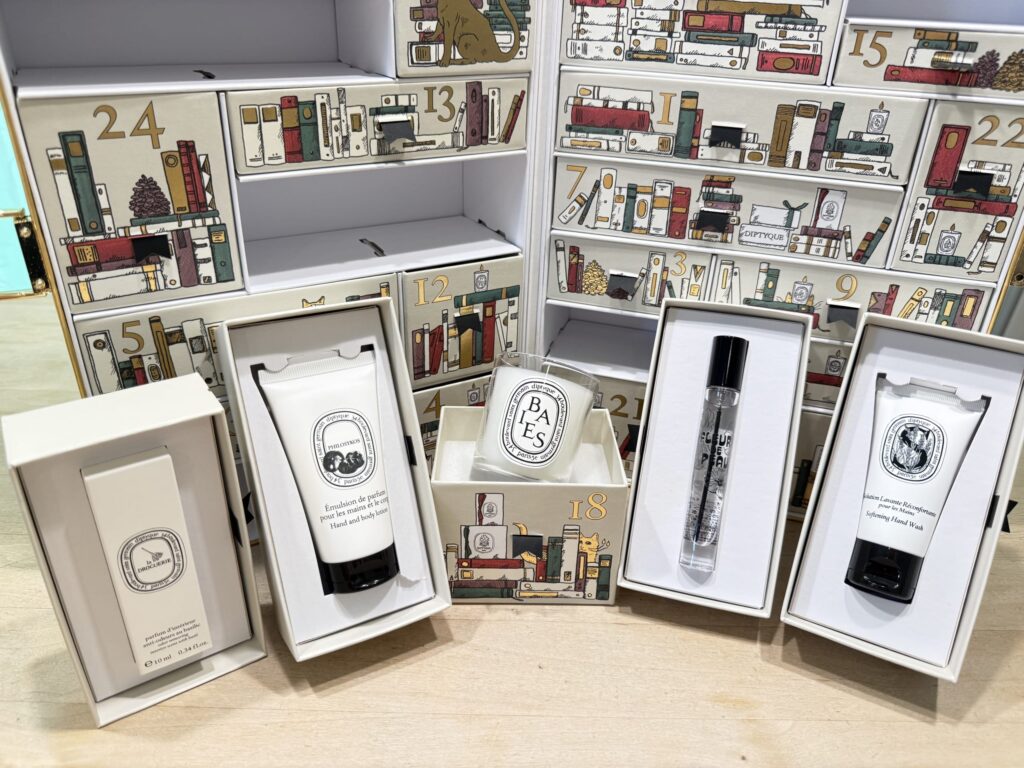A Diptyque advent calendar on a wooden table. In front of the drawers illustrated with bookshelves, five products from days 16 to 20 are displayed, including a mini candle, perfume, body lotion, and hand wash.