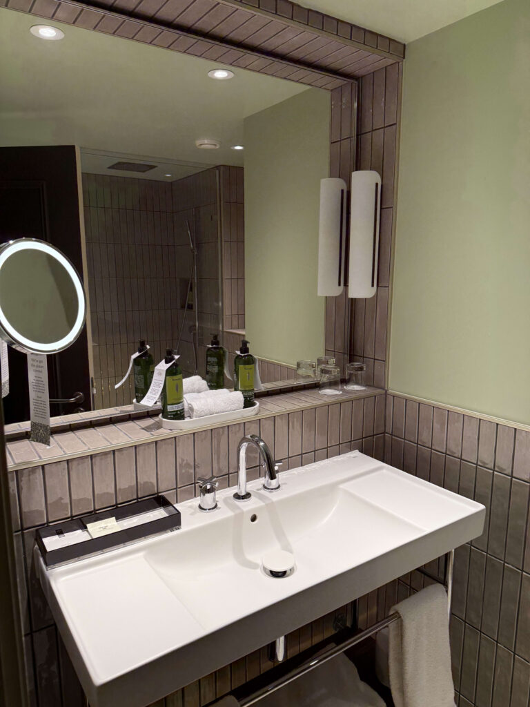 A modern hotel bathroom vanity featuring a white sink, rectangular wall tiles, a mirror, and a set of L'Occitane amenities.