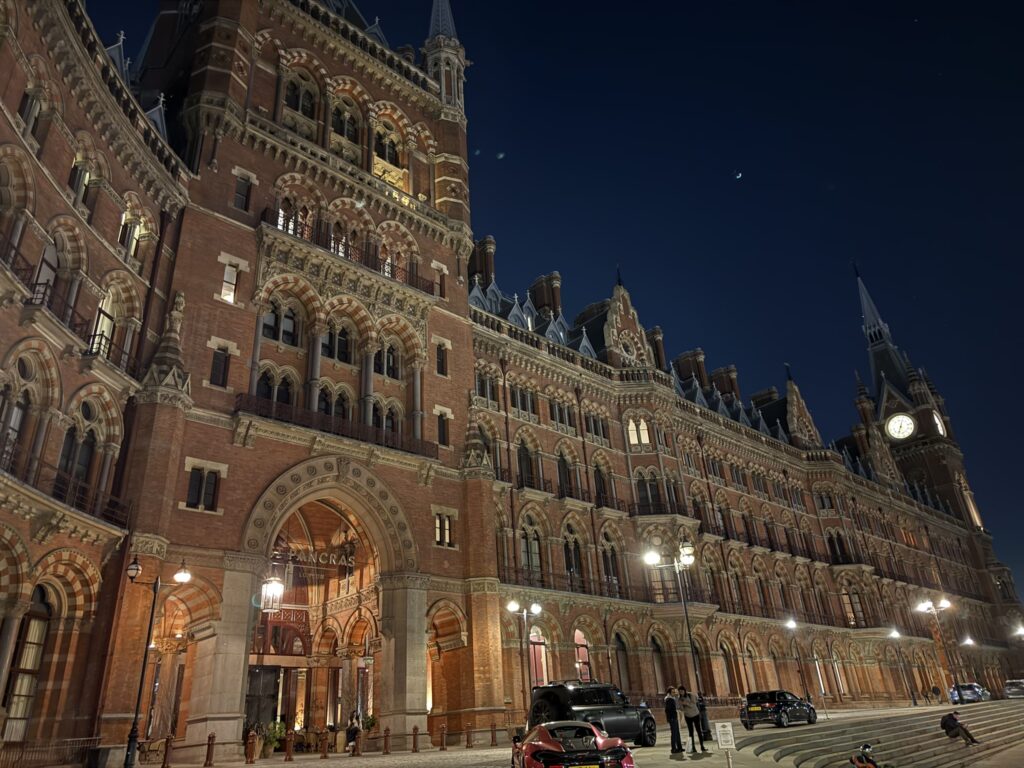 夜空の下、暖色の光に照らされたロンドンのセント・パンクラス駅の壮大なゴシック様式の赤レンガの建物。空には三日月が浮かんでいる。