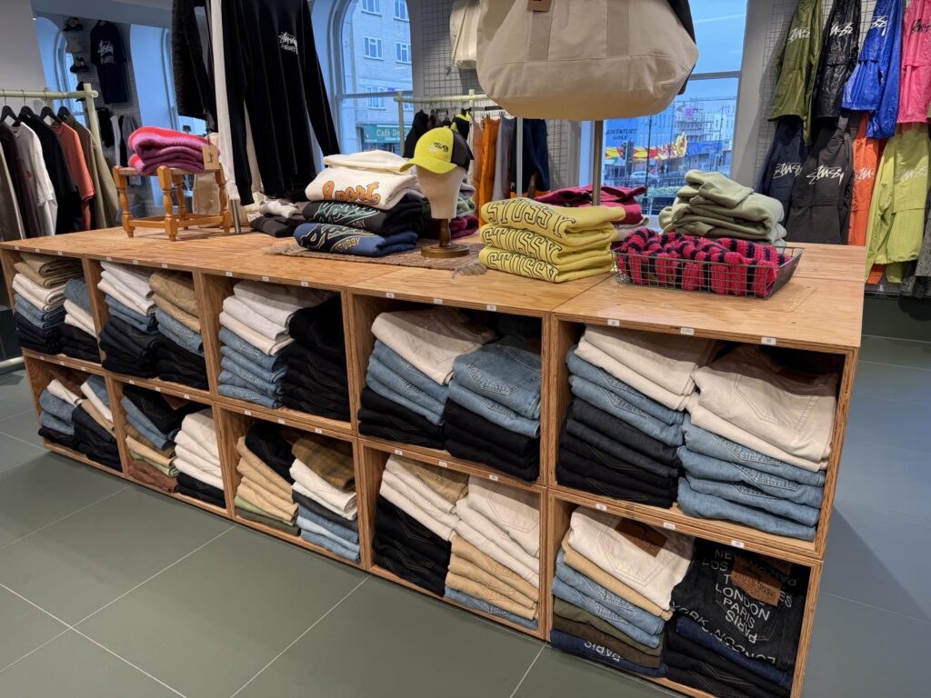Stussyの店舗内。木製の陳列棚に様々な色のデニムがきれいに折り畳まれて並べられている。棚の上にはスウェットや帽子も展示されている。