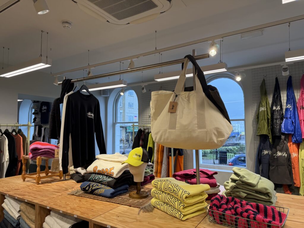 ストリートウェアブランドの店内で、木製の陳列台にカラフルなスウェットシャツや帽子、トートバッグが並べられている。背景にはハンガーにかかった服や窓が見える。