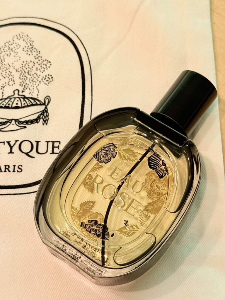 A close-up photo of a bottle of Diptyque's 'Eau Rose' perfume resting on a white drawstring pouch with the brand's logo. The glass bottle is decorated with gold and black rose illustrations.