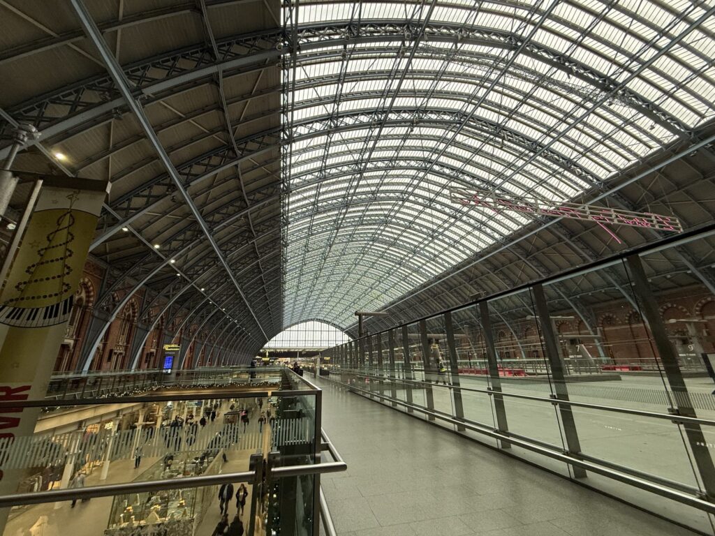 広大な駅の構内。2階の通路から、壮大なアーチ型の鉄骨とガラスの天井を見上げ、階下を行き交う人々や店舗を見下ろしている。