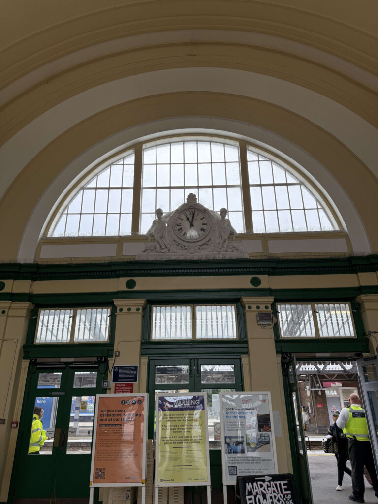 The interior of Margate station. Beneath a cream-colored arched ceiling, there is a large white clock with sculptures and an arched window. Below, green-painted doors and window frames are visible.