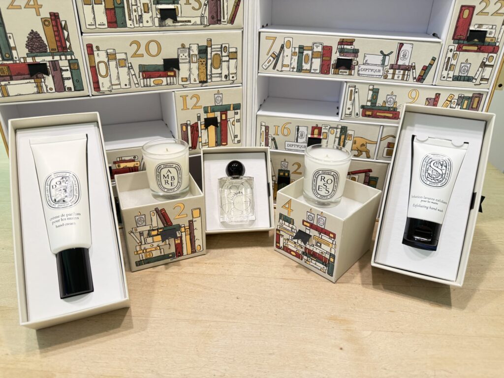 A Diptyque advent calendar on a wooden table. The boxes for days 1 through 5 are open, with the contents - two mini candles, a perfume, a hand cream, and a hand wash - displayed in front.