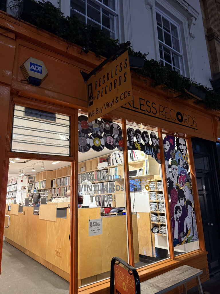 The storefront of Reckless Records, brightly lit with an orange facade at night. The windows are decorated with illustrations of musicians and vinyl records, and shelves of records are visible inside.