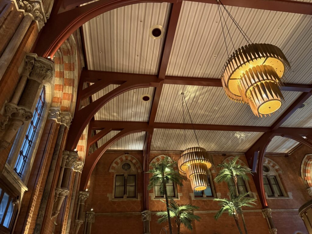 The interior of a building with red brick and stone walls and a high ceiling. Large wooden chandeliers hang from the striped ceiling crossed with red beams, casting a warm light. Palm trees are also visible inside.
