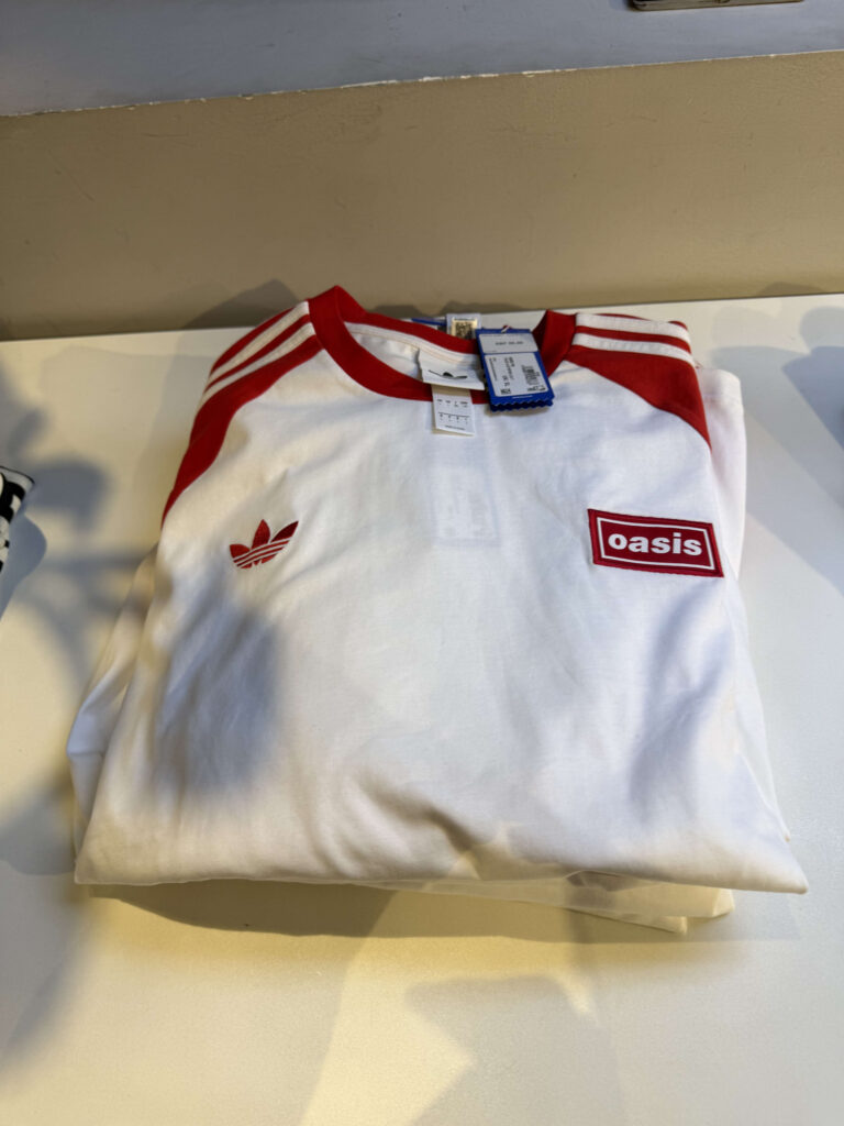 An Adidas x Oasis collaboration t-shirt is folded on a white shelf. The shirt is white with red accents, featuring the logos of both brands on the chest.