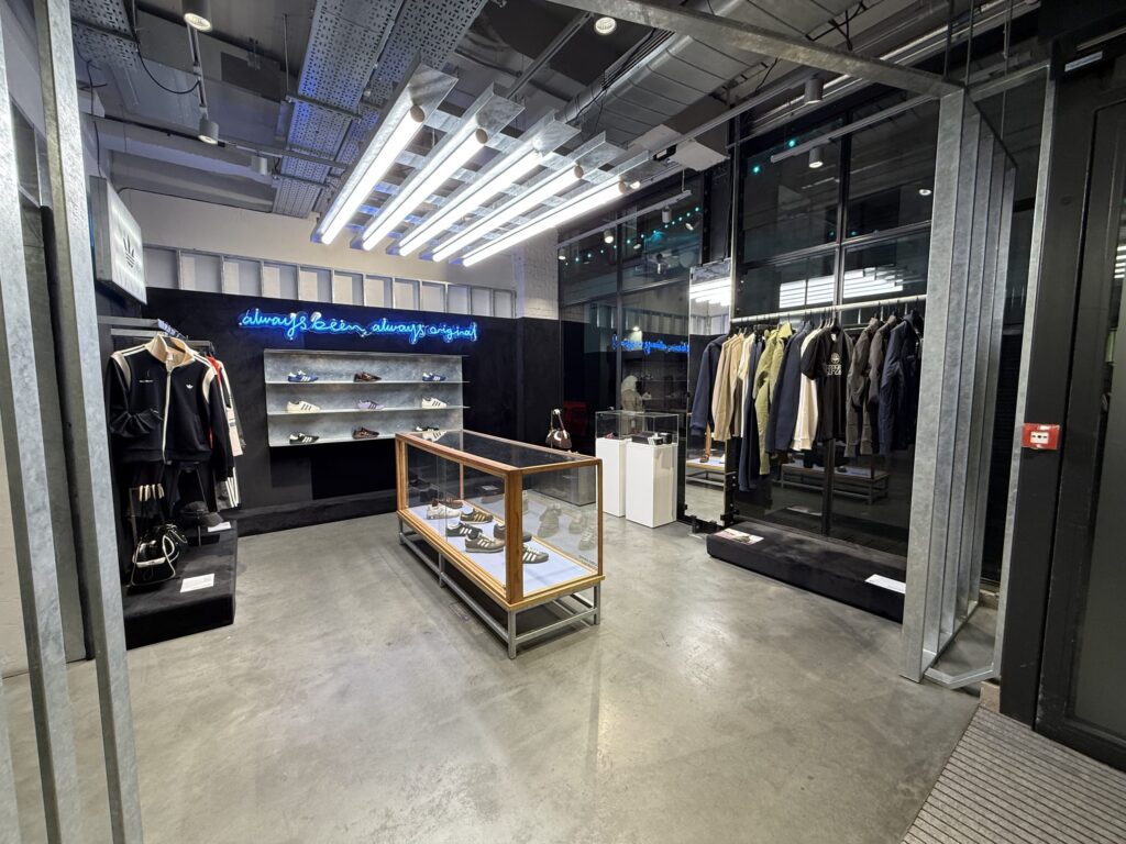 The interior of an apparel shop with an industrial design. It features a concrete floor, an exposed ceiling, sneakers displayed on shelves, a glass case, clothes on a rack, and a blue neon sign on the back wall.