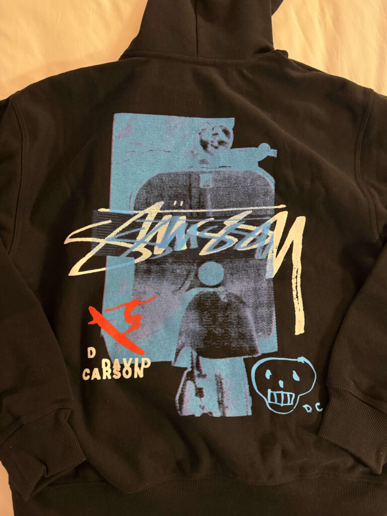 The back of a black hoodie from the Stüssy and David Carson collaboration. A large, white Stüssy logo is printed over a blue abstract graphic, accompanied by a red surfer illustration and a hand-drawn style skull.
