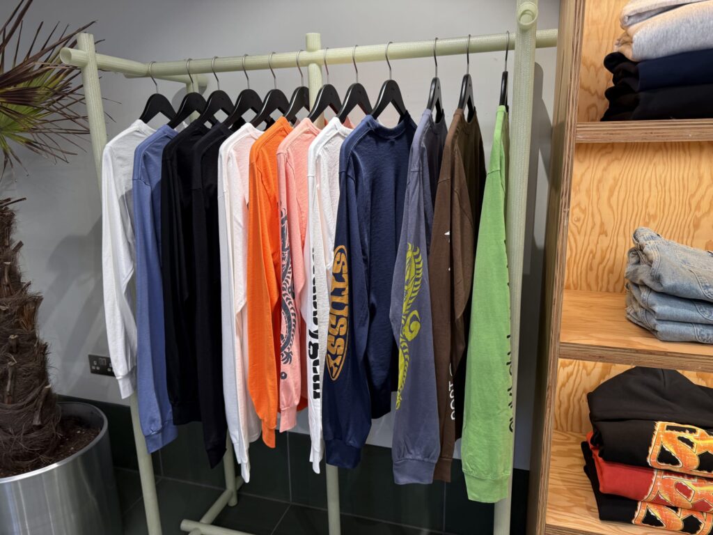 Long-sleeved T-shirts in various colors like white, blue, orange, and green hanging on a clothing rack inside a store. To the right is a wooden shelving unit with folded jeans and hoodies.