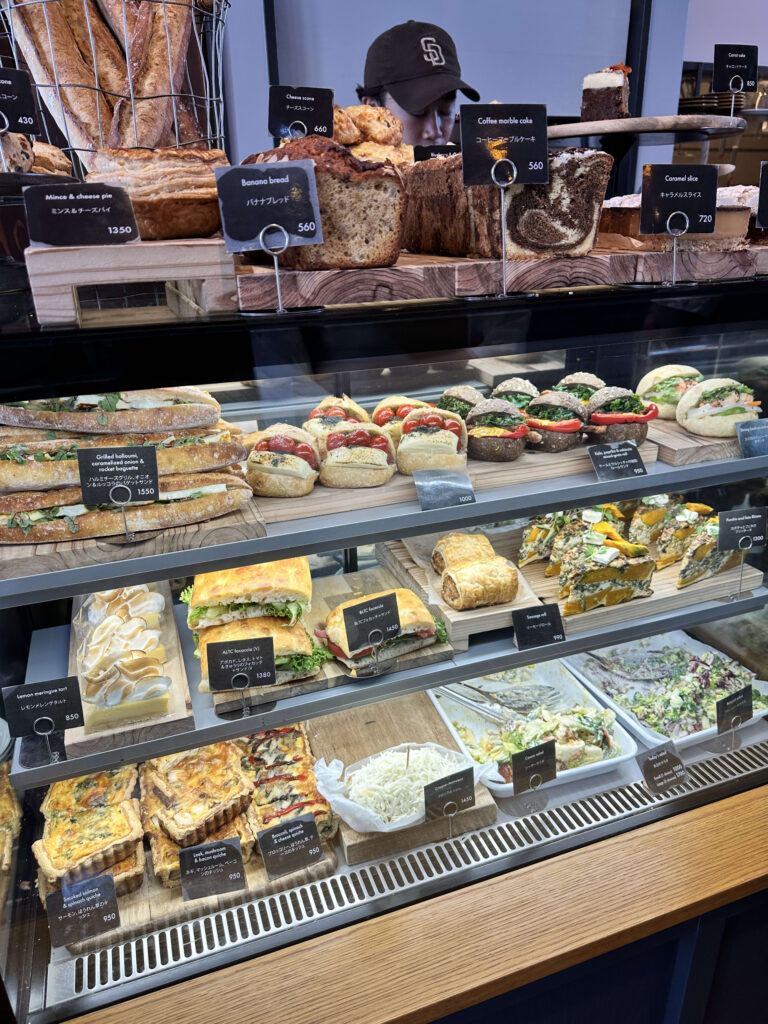 ベーカリーのガラス製ショーケースに、パン、ケーキ、サンドイッチ、キッシュ、サラダなど、美味しそうな商品が種類豊富に陳列されている様子。