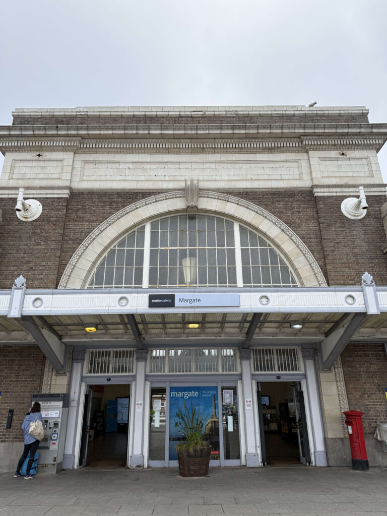 The front entrance of Margate station in the UK under an overcast sky. The classic station building is made of brown brick and white stone, with a large arched window above the entrance.