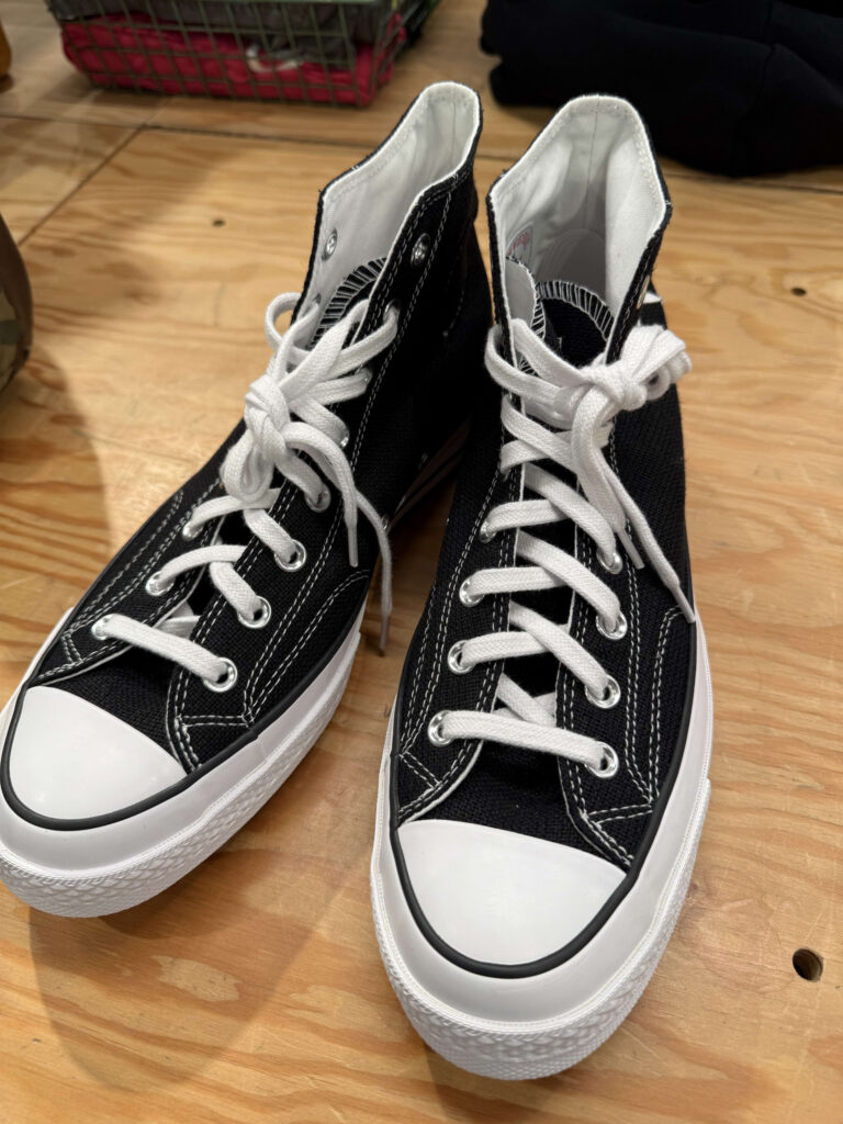 A pair of black high-top collaboration sneakers with Converse, featuring white laces and toe caps, placed on a wooden table.