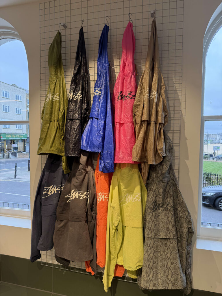 Various colorful Stüssy jackets hanging in two rows on a wire mesh rack against a wall in a store.
