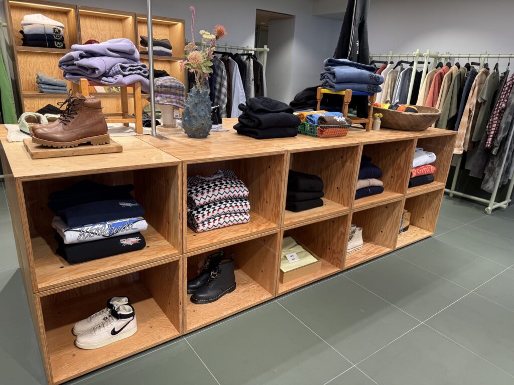 衣料品店の店内。中央にある木製の陳列棚には、折りたたまれた洋服や靴がディスプレイされている。背景には洋服ラックが並んでいる。