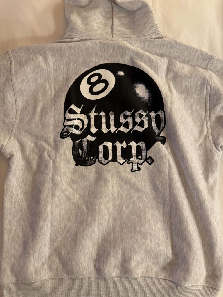 ライトグレーのフーディーの背面。中央に黒いエイトボールと、その上にゴシック体の白文字で「Stüssy Corp.」と書かれたグラフィックがプリントされている。