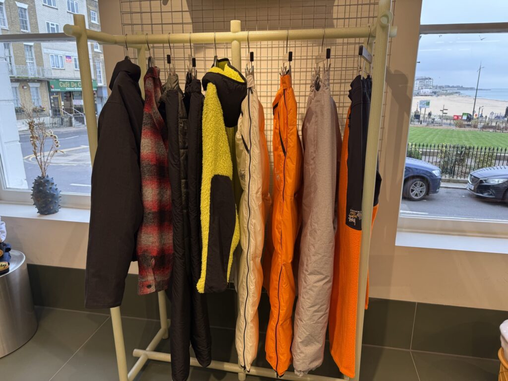 淡い緑色の洋服ラックに、色とりどりのジャケットやパンツが掛けられている。窓の外には海辺の街並みが見える。