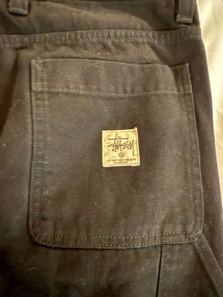 Stüssyの黒いワークパンツのバックポケットのクローズアップ。中央にはStüssyのロゴと「AUTHENTIC WORKGEAR」の文字が入ったタグが付いている。