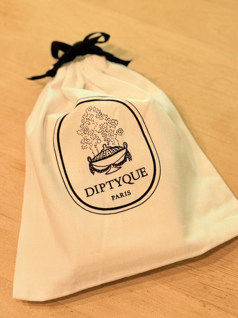 木製のテーブルの上に置かれた、黒いリボンで結ばれたディプティックのロゴ入り巾着ポーチ。